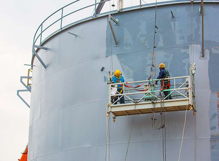 Workers inspecting a large industrial storage tank on suspended scaffolding.