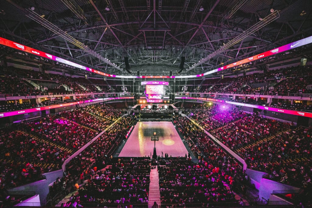 Spectator-filled basketball arena illuminated with pink and purple lighting, large LED displays.