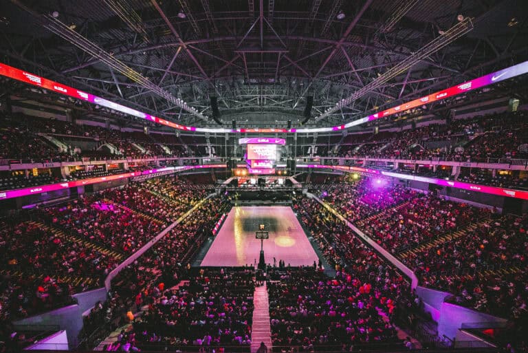 Spectator-filled basketball arena illuminated with pink and purple lighting, large LED displays.