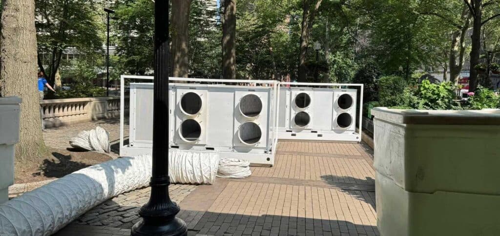 Road construction barriers with speakers blocking a park walkway, surrounded by trees and urban elements.