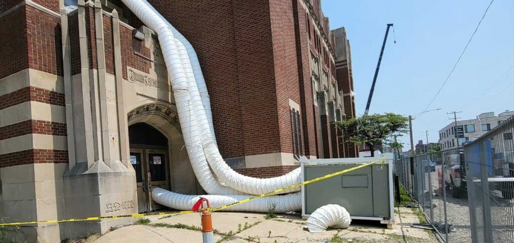 Damaged building exterior with large ventilation ducts, construction barrier, and fencing, indicating ongoing construction or repair work.