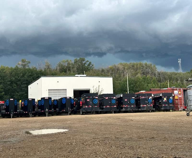 Row of black heavy equipment trailers parked outside a warehouse under stormy skies.