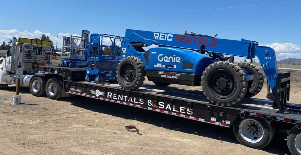 REIC Rentals blue Genie telehandler on flatbed trailer at construction site.